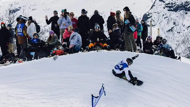 Snowboarder kjører gjennom en sving i løypa mens publikum sitter og ser på fra kanten av bakken.
