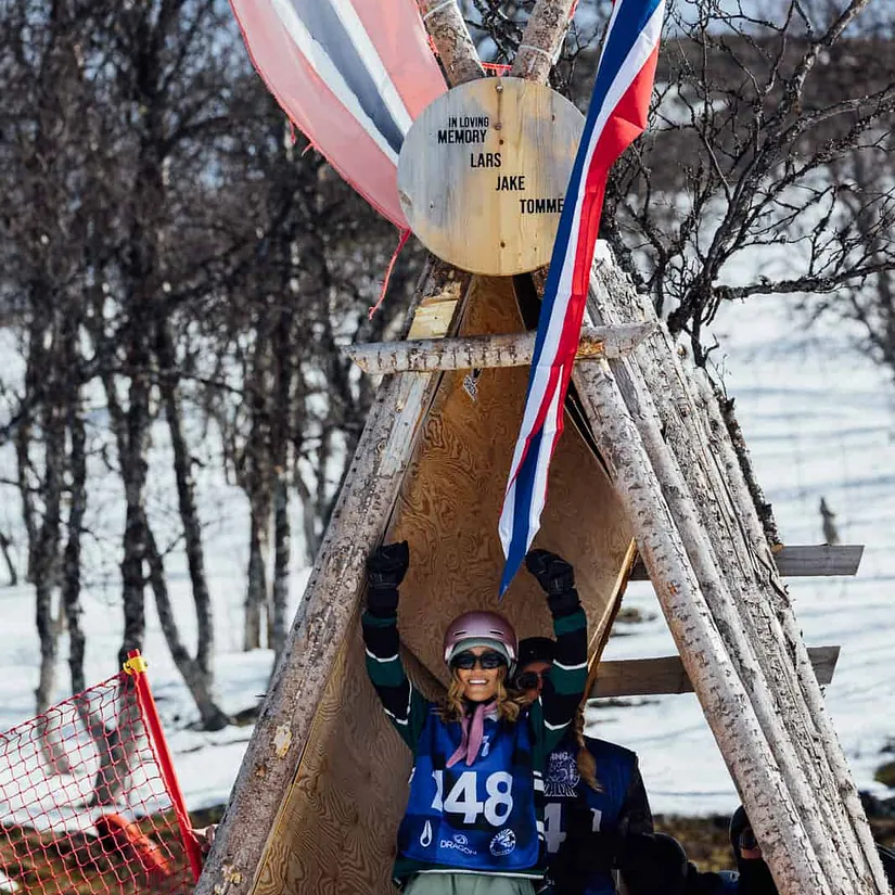Minnemarkering med norske flagg og trekonstruksjon i snødekt fjellterreng under arrangement.