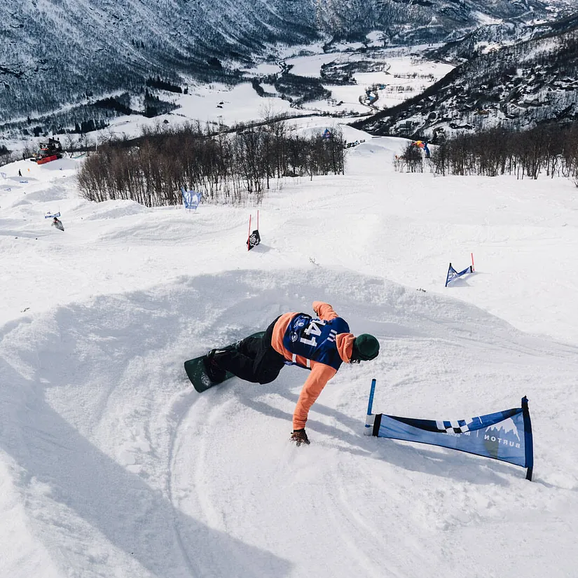 Snowboarder carver gjennom en sving i løypa med snødekte fjell og dal i bakgrunnen.